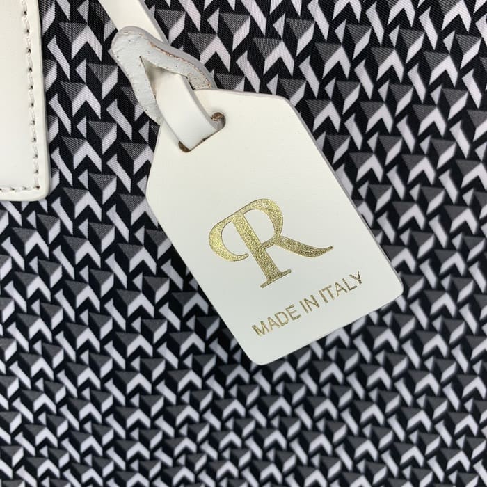 ป้ายหนัง ROBERTA PIERI สีขาวที่มีตัวอักษร "R" เป็นสีทองและคําว่า "MADE IN ITALY" ห้อยลงมาจากพื้นหลังของผ้าเรขาคณิตขาวดํา
