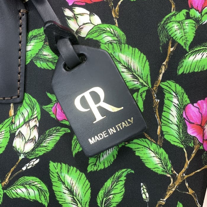 ป้ายติดกระเป๋า ROBERTA PIERI สีดําที่มีตัวอักษร "R" สีทองและข้อความภาษาอิตาลีแขวนอยู่บนผ้าลายดอกไม้ที่มีใบไม้สีเขียวและดอกไม้สีชมพู
