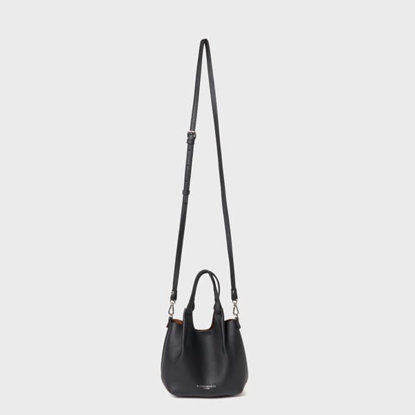 กระเป๋าถือ Gianni Chiarini Dua XS หนังสีดํา มีด้ามจับสั้นและสายรัดสะพายข้างปรับความยาวได้ ตั้งตรงตัดกับพื้นหลังสีเทาอ่อนธรรมดา